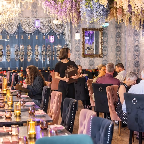 A restaurant dining room with patrons seated at tables, a server carrying menus, and floral decor.