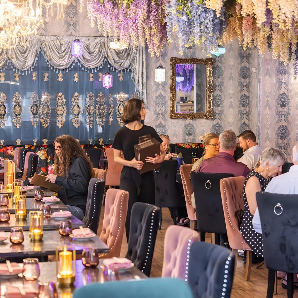 A restaurant dining room with patrons seated at tables, a server carrying menus, and floral decor.