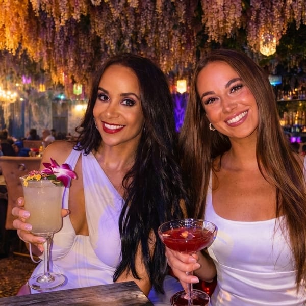 Two women in white dresses holding cocktails in a restaurant with hanging floral decor.