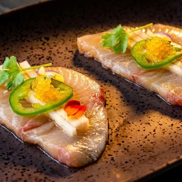 Three pieces of sashimi with jalapeño, fish roe, and herbs on a dark speckled plate.