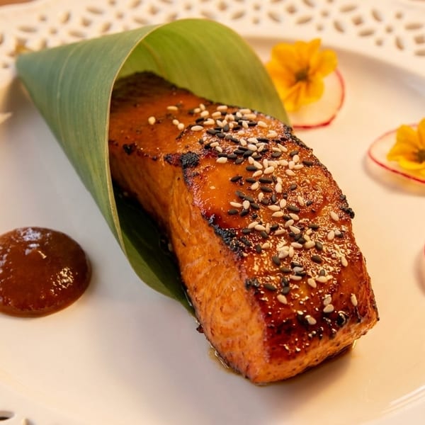 Glazed salmon with sesame seeds, wrapped in a banana leaf, with sauce and radish garnishes.