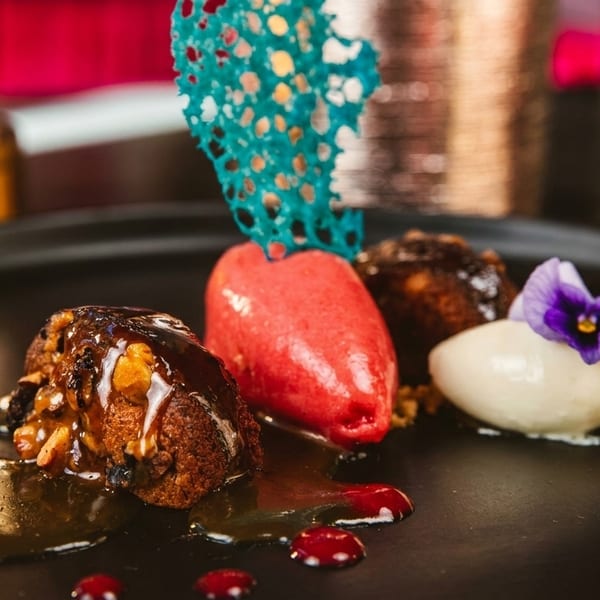 Gourmet dessert with cake, red sorbet, white ice cream, and a purple flower on a black plate.