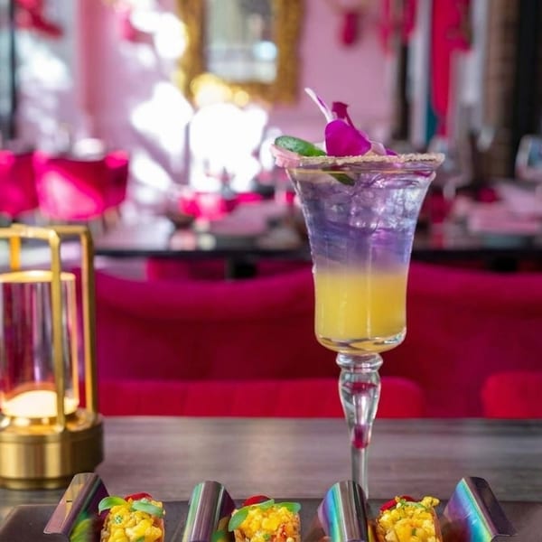 A vibrant purple and yellow cocktail with appetizers and a golden lantern on a table in a pink restaurant.