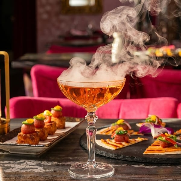 Smoking cocktail with appetizers on a dark table, pink chairs in the background.